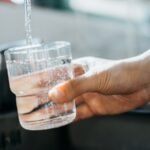 A stock image of a person holding a glass under a running faucet. The cup is filling with drinking water.
